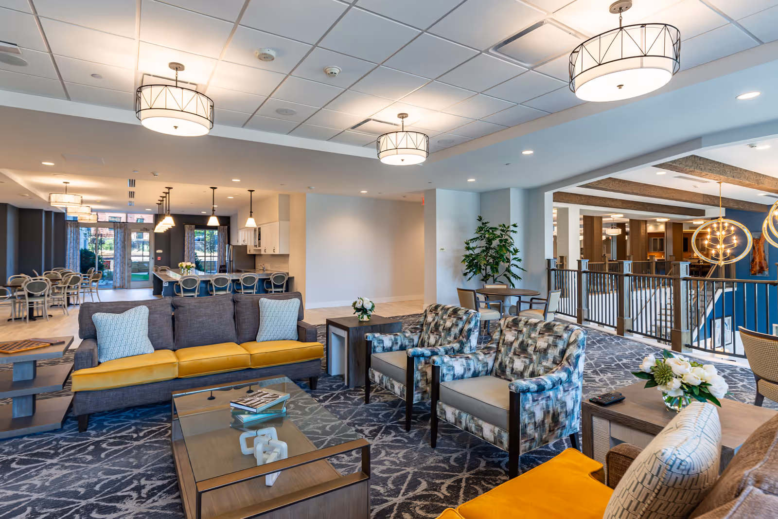 A spacious and well-lit living room area in a senior living facility featuring a gray sofa with yellow cushions, two patterned armchairs, a glass coffee table with books, and a side table with a flower arrangement. In the background, there is a dining area with multiple tables and chairs, pendant lighting, and large windows letting in natural light.