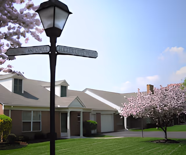 Single-story senior living building front with a lamp post, manicured lawn, and blooming pink trees.