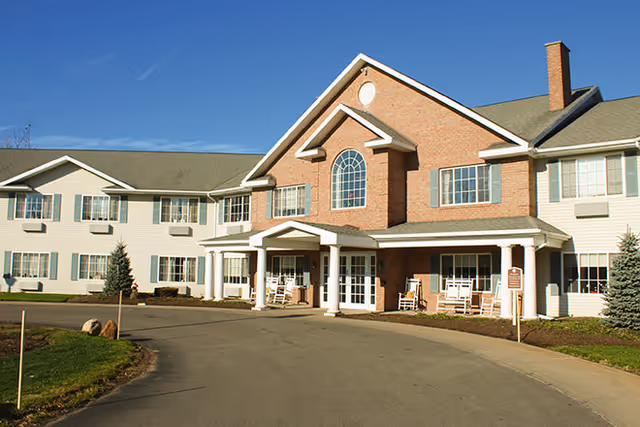 Exterior front view of a two-story senior living facility building with a brick and white siding facade, multiple windows, a covered entrance with white columns, and rocking chairs on the porch under a clear blue sky.