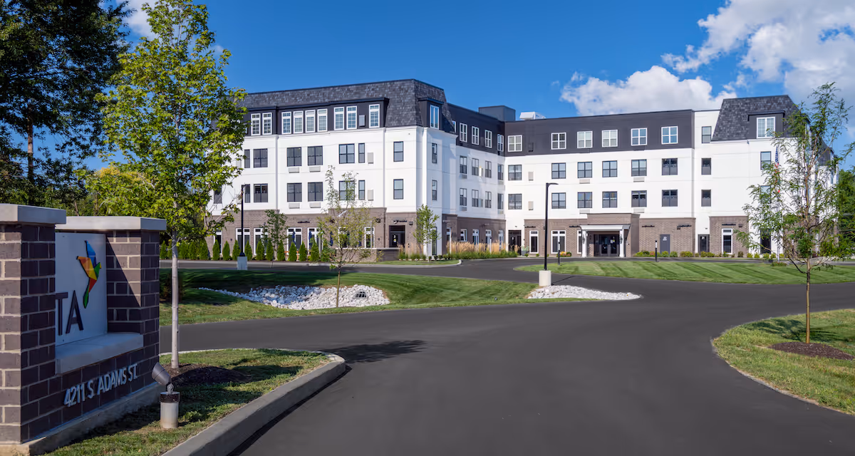 Entrance view of the Vita of Marion senior living building: a four-story white-and-gray facility with a circular driveway, lawn, and a sign at the front.
