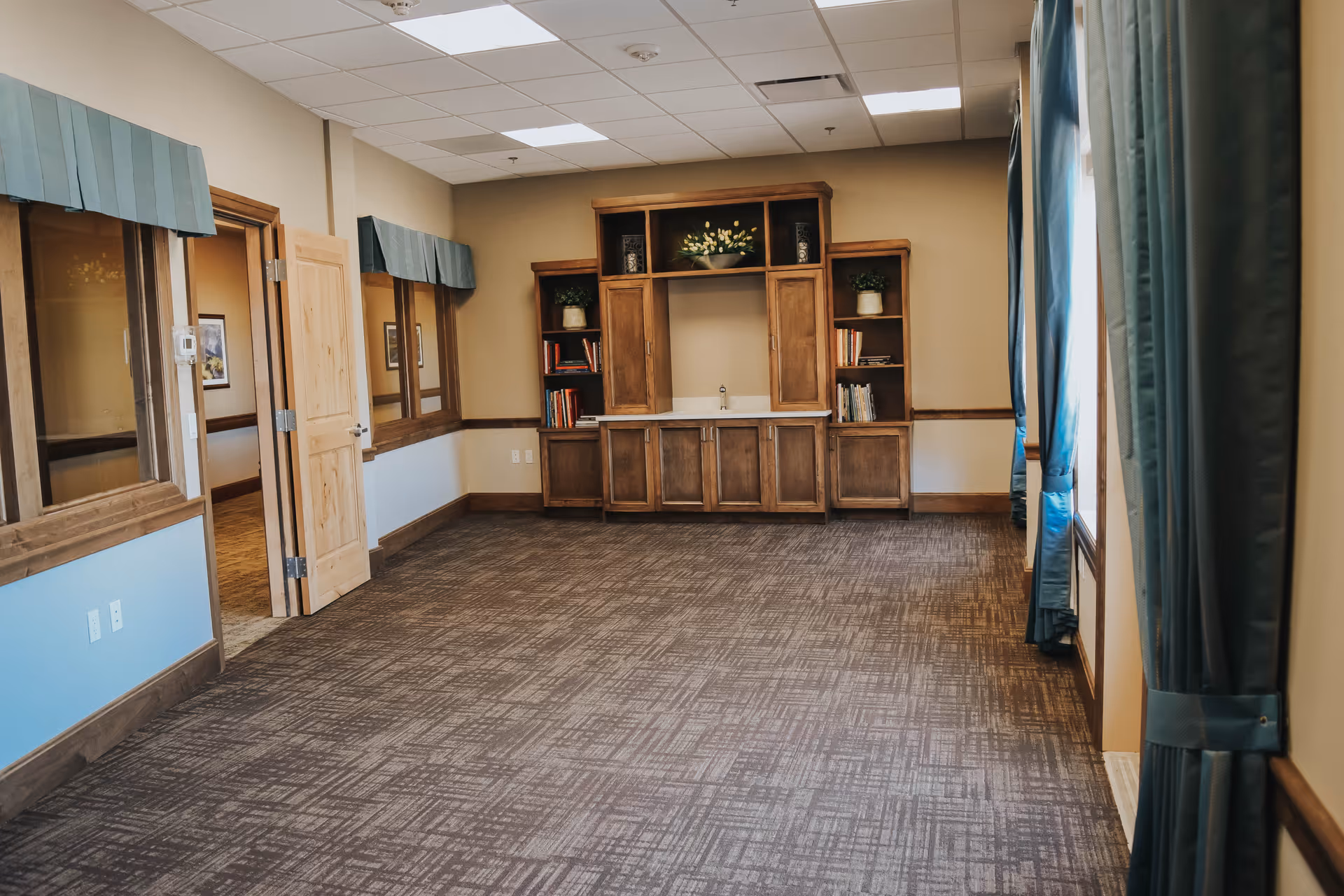 Carpeted empty community room with wooden built-in cabinetry and a small sink, windows with teal curtains, and an open doorway.