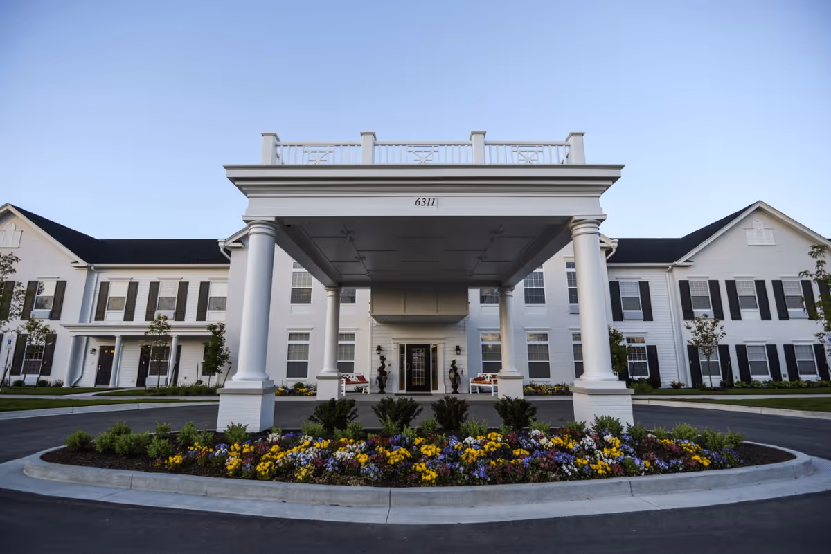 Front exterior view of a large white senior living facility building with black shutters, a covered entrance supported by four white columns, and a landscaped flower bed with colorful flowers in front.