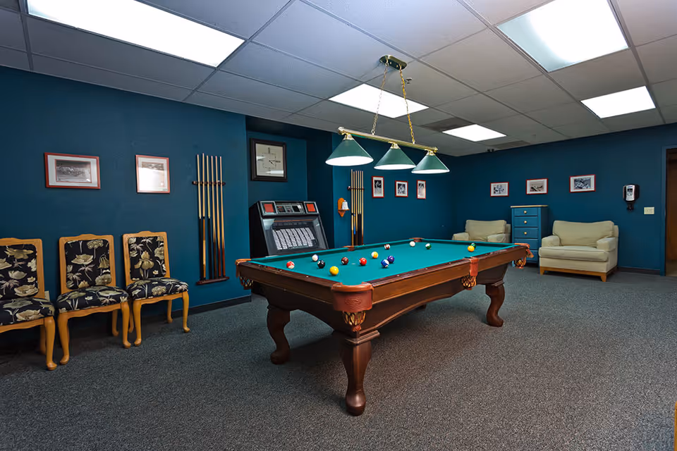 A recreational room with a pool table in the center, surrounded by several pool cues mounted on the wall. There are three floral-patterned chairs on the left side and two beige armchairs with a blue chest of drawers between them on the right side. The walls are painted dark teal and decorated with framed pictures. The ceiling has recessed lighting and a hanging light fixture with three green lampshades above the pool table.