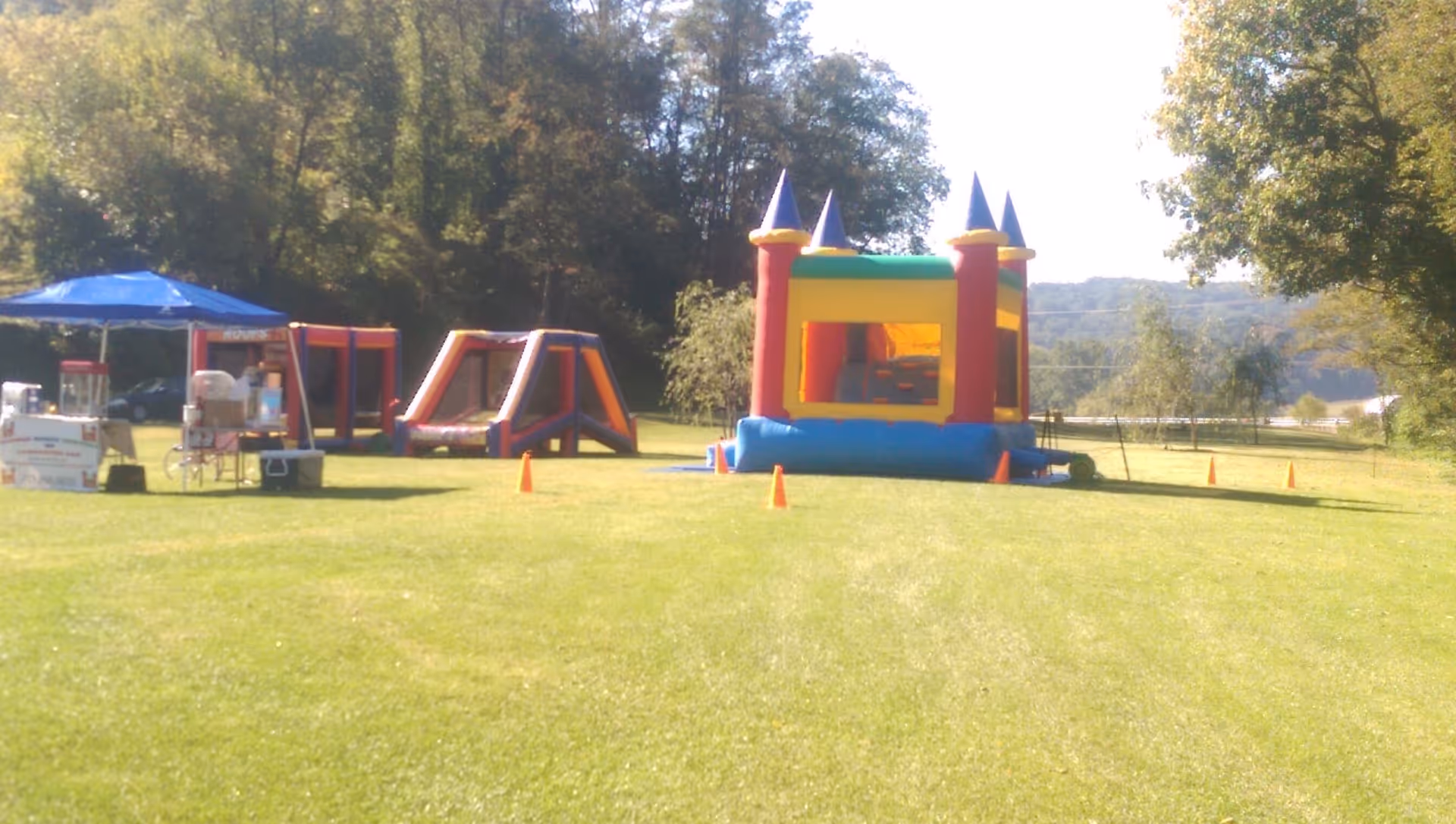 Outdoor grassy area with inflatable bounce house and inflatable obstacle course set up, surrounded by orange cones. There is a blue canopy tent with tables and equipment underneath on the left side. Trees and hills are visible in the background under a sunny sky.