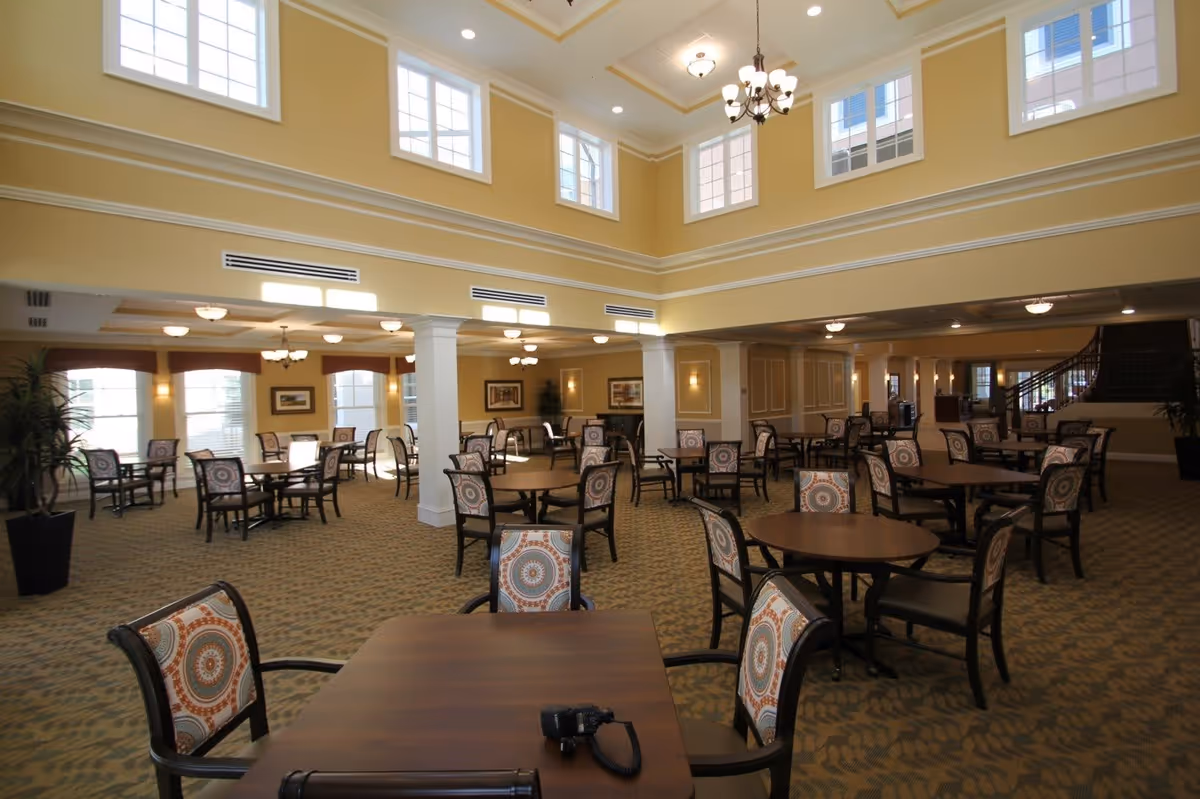 Spacious dining room with multiple round wooden tables and patterned cushioned chairs arranged neatly. The room features high ceilings with large windows near the top, allowing natural light to fill the space. There are decorative columns, chandeliers, and framed artwork on the walls. The carpet has a subtle pattern, and a staircase is visible in the background.