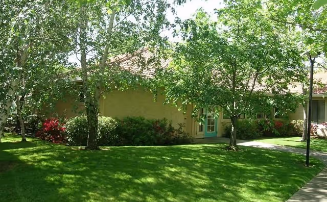 Single-story apartment building entrance partially obscured by trees with a manicured lawn and walkway.