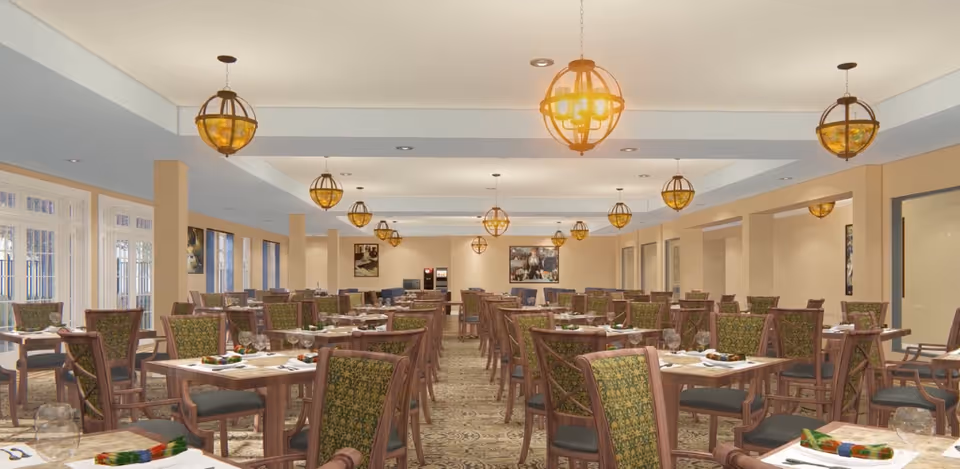 A spacious dining room with multiple wooden tables and chairs arranged neatly. Each table is set with plates, glasses, and colorful napkins. The room features large windows on the left side allowing natural light to enter, and several decorative hanging light fixtures on the ceiling. The walls are painted a light beige color and there are framed pictures hanging on the far wall.