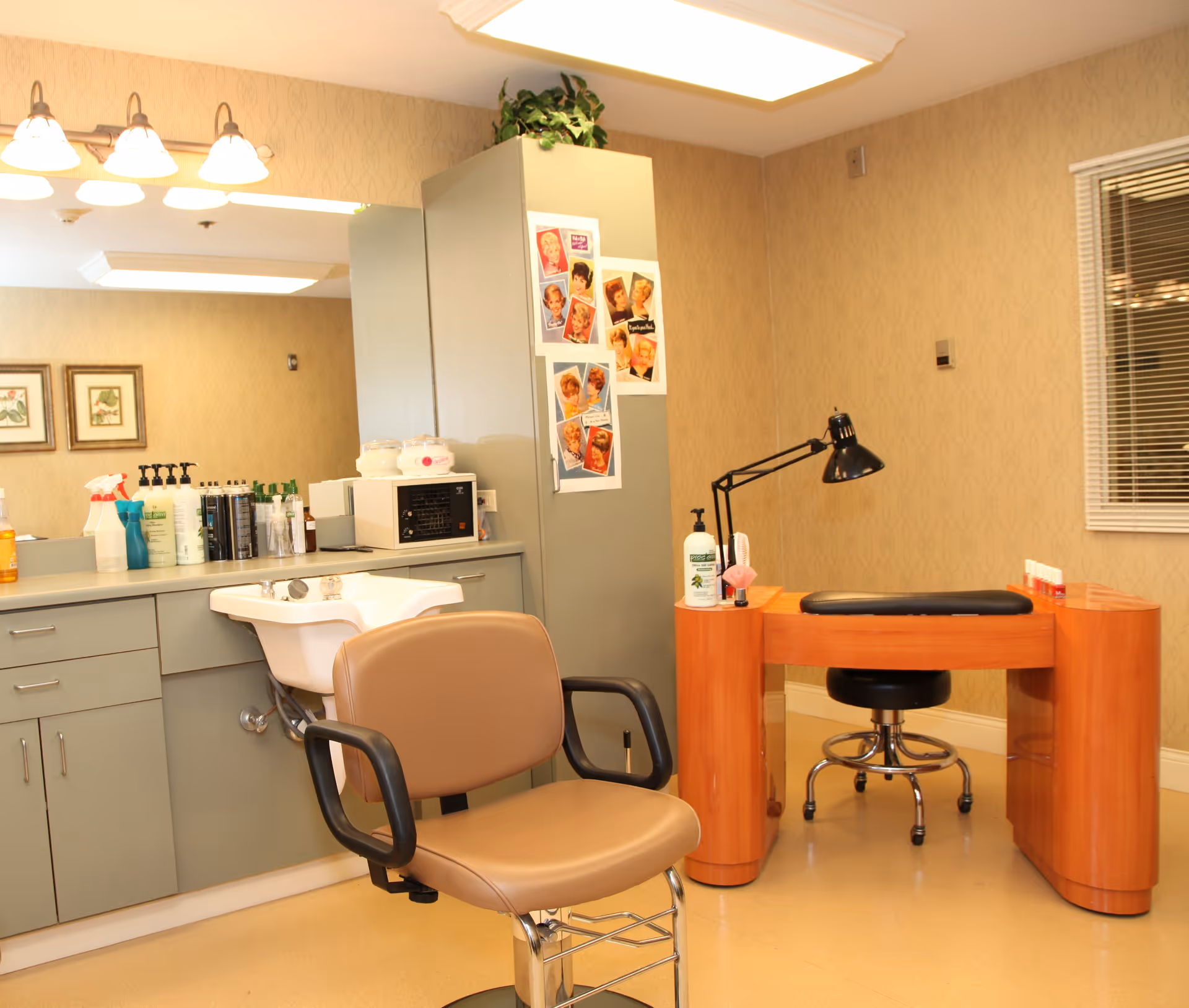 Interior view of a salon area in a senior living facility featuring a beige salon chair in front of a white sink, a countertop with various hair and beauty products, a wooden manicure table with a black adjustable lamp, and a black stool. The walls are decorated with light patterned wallpaper and a window with blinds is visible on the right side.
