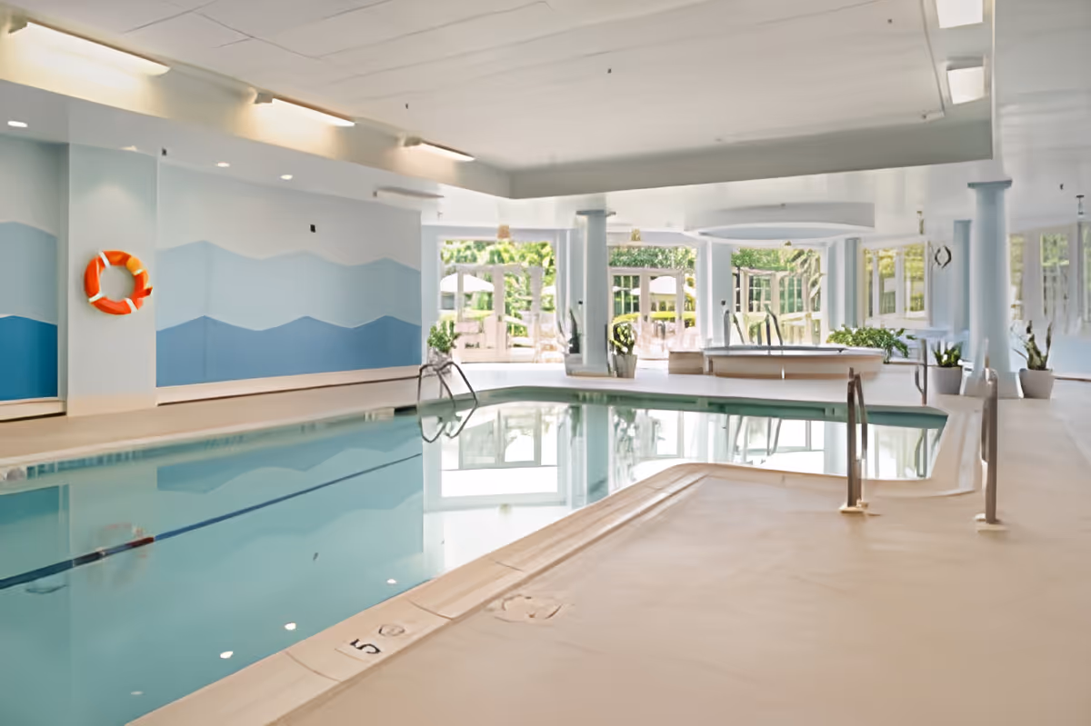 Indoor swimming pool area with light blue and white walls featuring wave patterns, a life preserver mounted on the wall, pool ladders, and large windows letting in natural light. There are plants and seating visible near the windows.