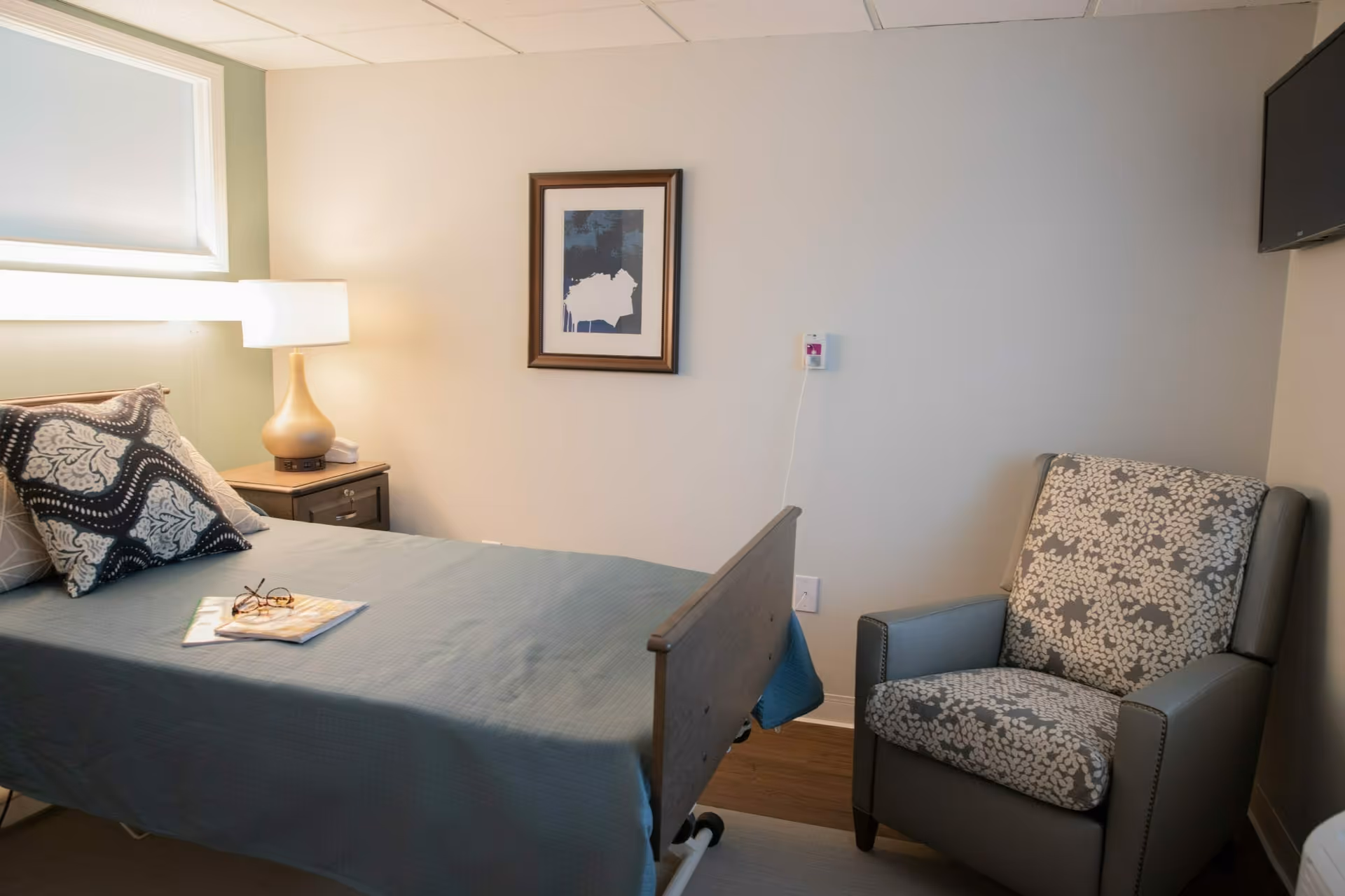 A simple resident bedroom with a single bed, bedside table and lamp, framed artwork on the wall, and an upholstered armchair.