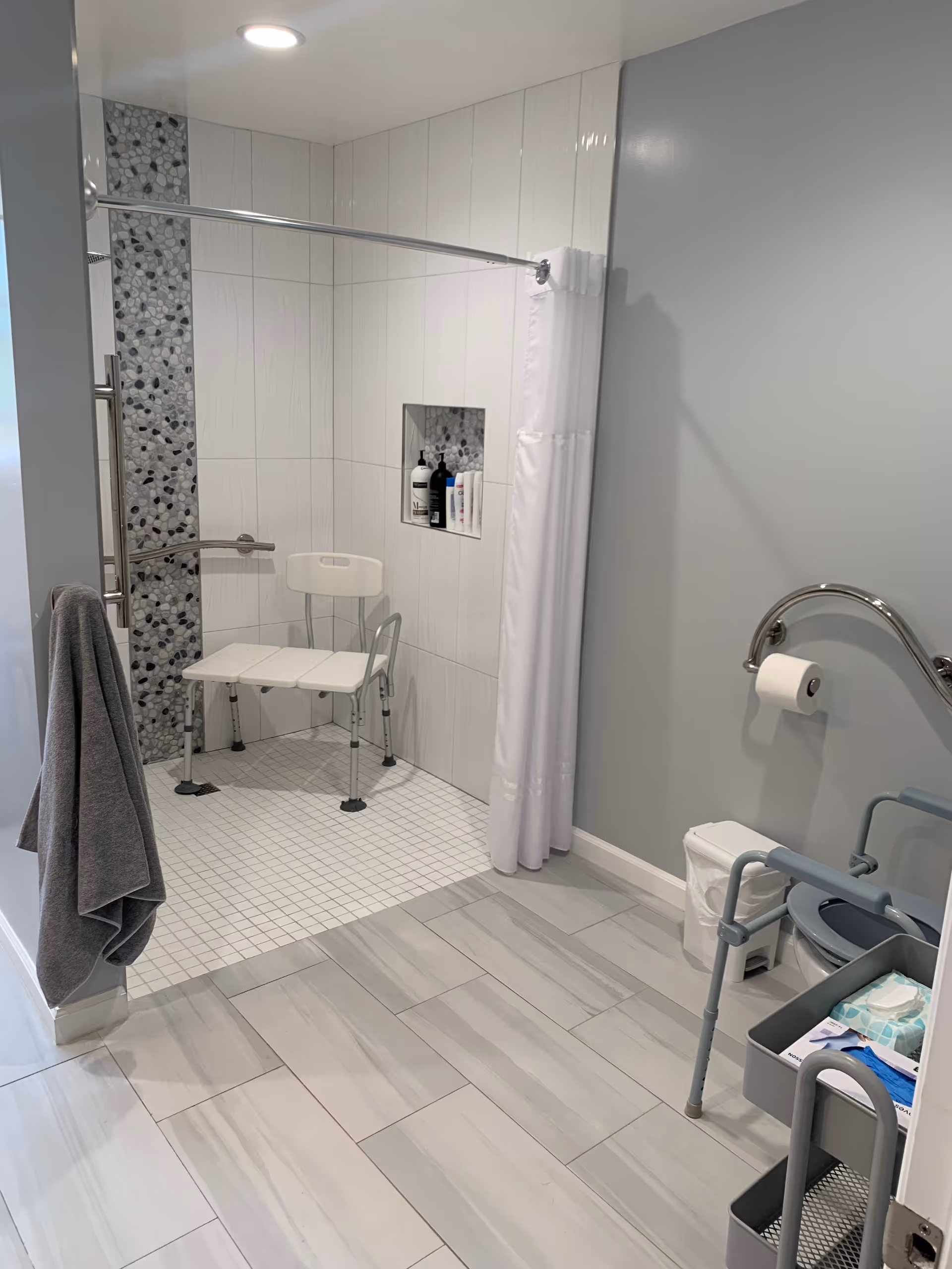 A modern accessible bathroom with a walk-in shower featuring a white shower chair, grab bars, and a white shower curtain. The shower wall has a vertical strip of pebble-like tiles. There is a gray towel hanging on a hook outside the shower. The bathroom floor is tiled in light gray, and there is a toilet with a grab bar and a small cart holding supplies nearby.