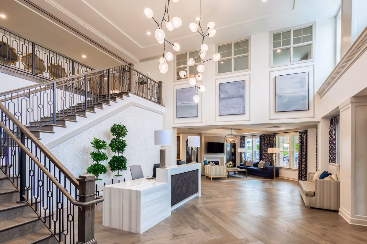 Spacious and elegant senior living facility lobby with a modern white reception desk, decorative plants, a large staircase with dark wood and metal railing, contemporary chandelier lighting, and a comfortable seating area with sofas and armchairs near large windows with patterned curtains.