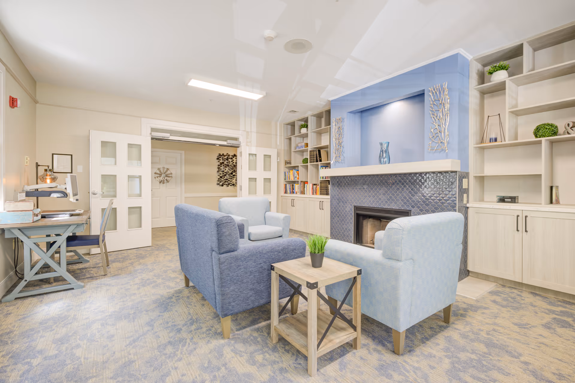 A bright common lounge with blue upholstered armchairs around a small wooden table facing a decorative fireplace and built-in shelving.