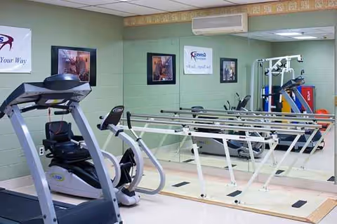 Indoor exercise room with treadmill, stationary bike, and parallel bars in a senior facility.