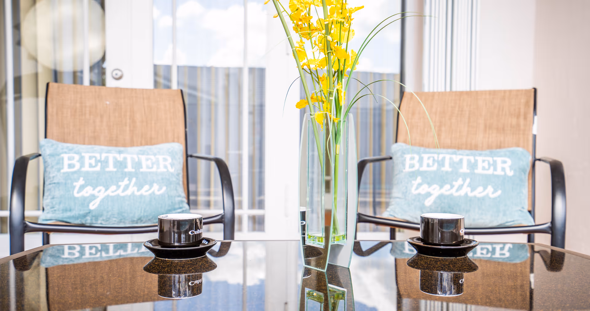 Two brown chairs with light blue pillows that say 'BETTER together' are positioned behind a glass table. On the table, there are two black coffee cups with saucers and a tall glass vase with yellow flowers. The background shows a glass door with vertical blinds.