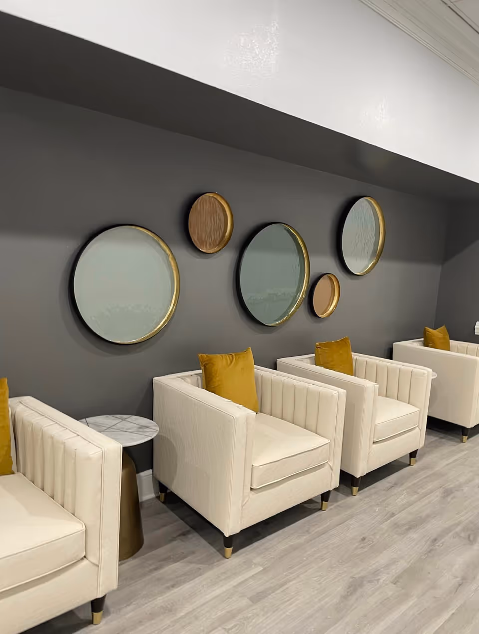 A row of cream-colored armchairs with mustard yellow pillows arranged against a dark gray wall. Above the chairs are decorative round mirrors with gold and black frames in various sizes. A small round side table with a marble top and gold base is placed between two of the chairs. The floor is light wood.