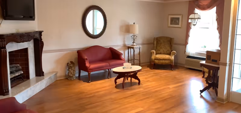 A cozy sitting room with hardwood floors featuring a fireplace, red loveseat, upholstered armchair, round coffee table, wall mirror and a window with drapes.