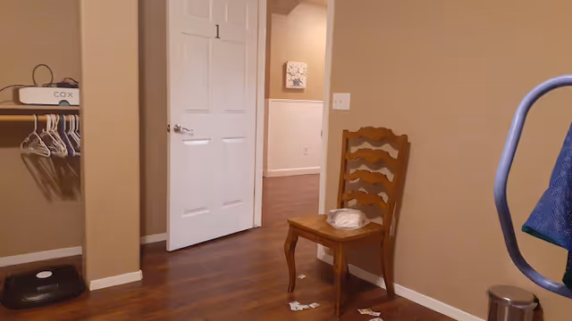 Interior view of a room with wooden flooring and beige walls. A wooden chair with a plastic bag on the seat is placed against the wall. There is an open closet on the left side with hangers and a Cox modem on the shelf. A white door is open, leading to a hallway with a framed picture on the wall. Part of a blue fabric item and a metal trash can are visible on the right side.