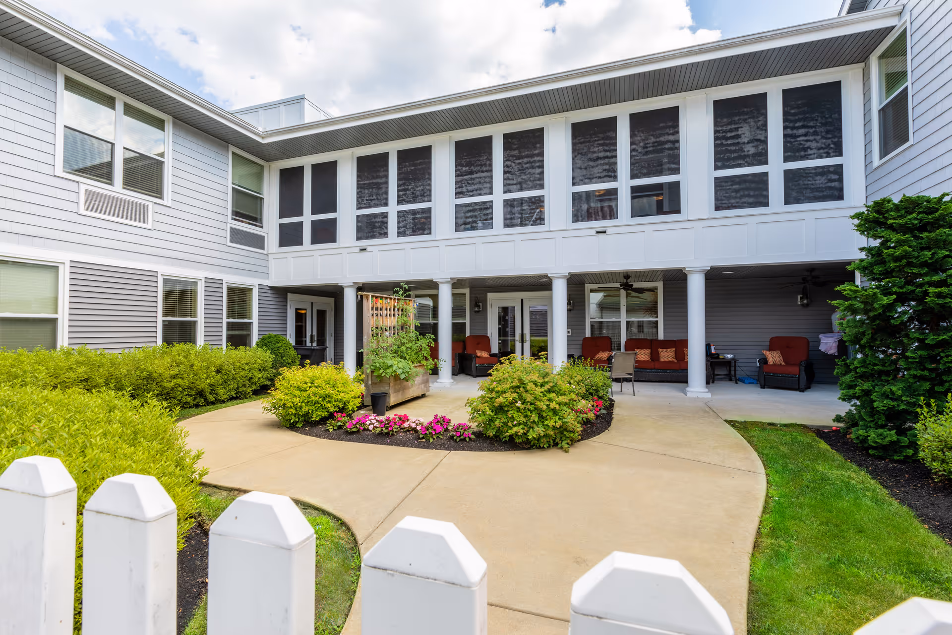 Outdoor courtyard area of a senior living facility with a paved walkway, green bushes, colorful flowers, and a white picket fence in the foreground. The building has gray siding, multiple windows, and a covered patio with seating including red cushioned chairs and a ceiling fan.
