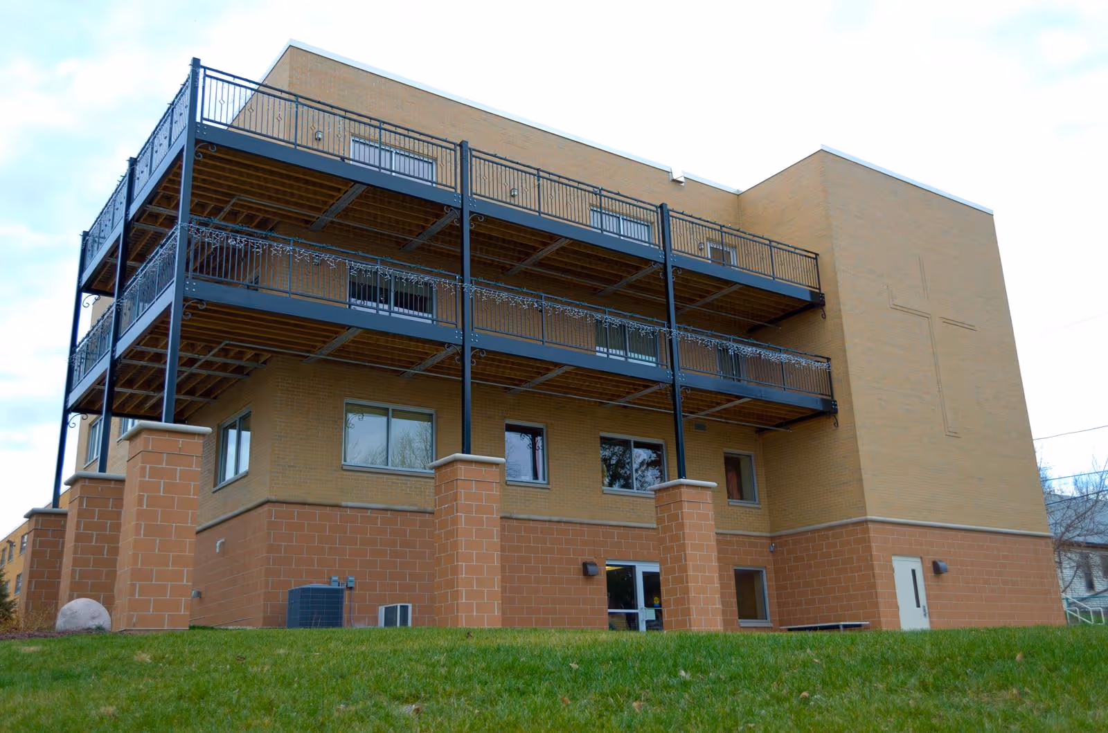 Exterior view of a multi-story brick building with balconies and a large cross design on one wall, surrounded by a grassy area under a cloudy sky.