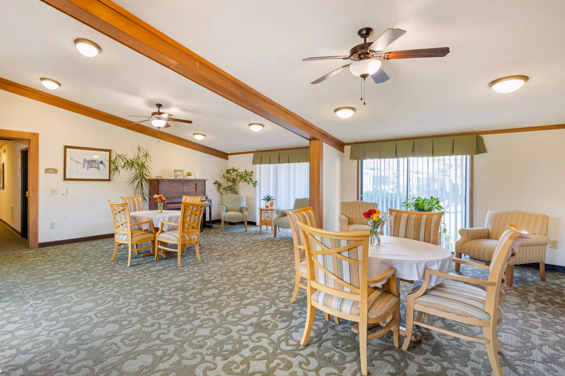 A bright and spacious common area with carpeted floor and wooden trim. The room features two round tables with chairs around them, each table decorated with a small vase of flowers. There are two ceiling fans with lights, large windows with green valances allowing natural light to enter, and several armchairs placed near the windows. A piano and some plants are visible in the background, creating a welcoming and comfortable atmosphere.