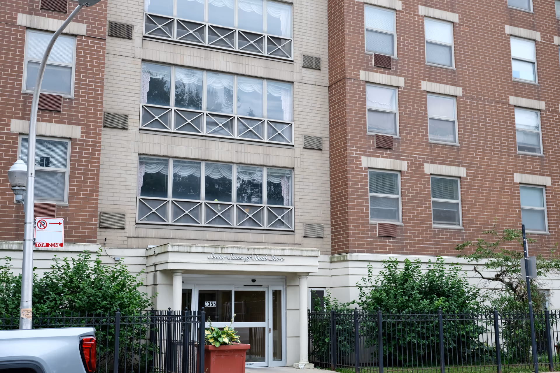 Exterior view of a multi-story senior living facility named Senior Suites of South Shore, showing the main entrance with glass doors, surrounded by brick and beige walls, windows with curtains, some greenery, a black metal fence, and a street lamp with a no parking sign.