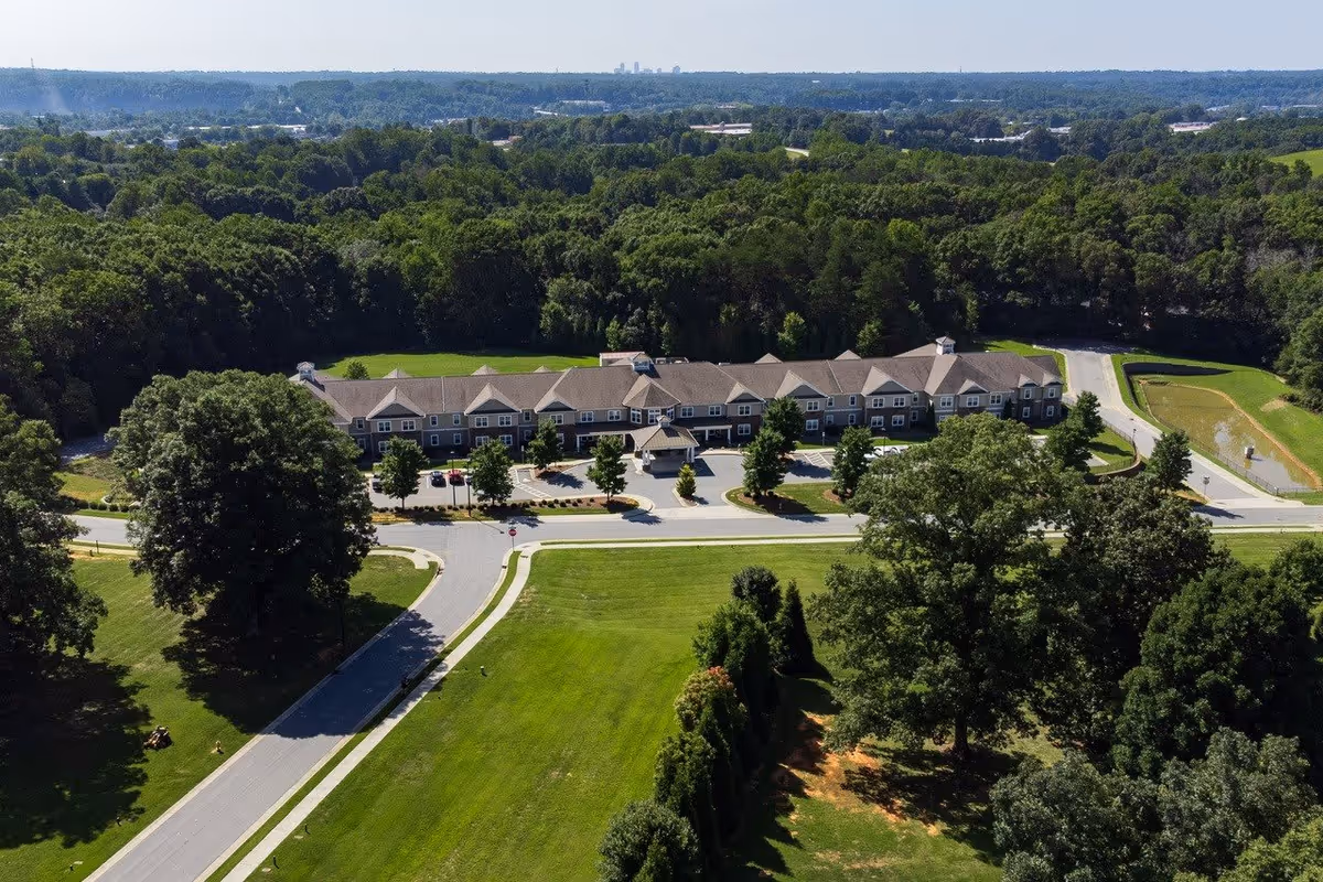 Aerial view of Arbor Ridge At Stanleyville facility surrounded by lush green trees and grass. The building is long with multiple peaked roofs and a parking area in front. Roads and landscaped areas are visible around the property.