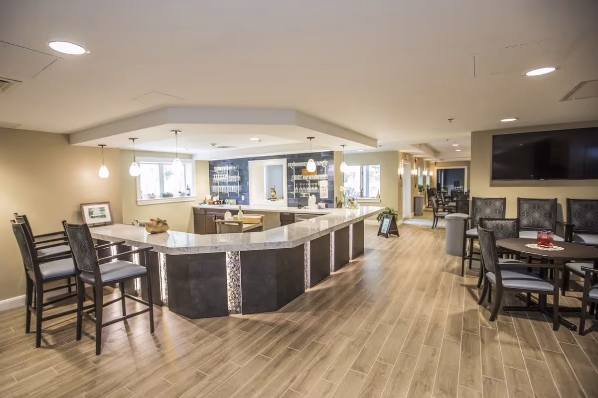 Interior view of a senior living facility lounge area with a large U-shaped counter with bar stools, pendant lights hanging above, and a seating area with tables and chairs. The space has wood-look flooring, beige walls, and a large TV mounted on the wall.