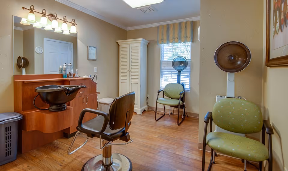 Interior of a hair salon area in a senior living facility with a styling chair in front of a mirror and sink, two green chairs, a hair dryer, a white cabinet, and a window with blinds and a valance.