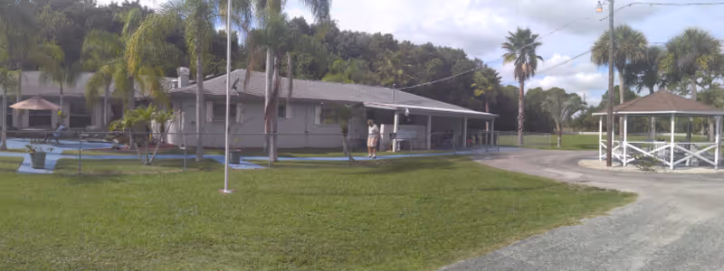 Single-story assisted living building with a covered porch, gazebo, palm trees, and a grassy lawn with a few people outside.