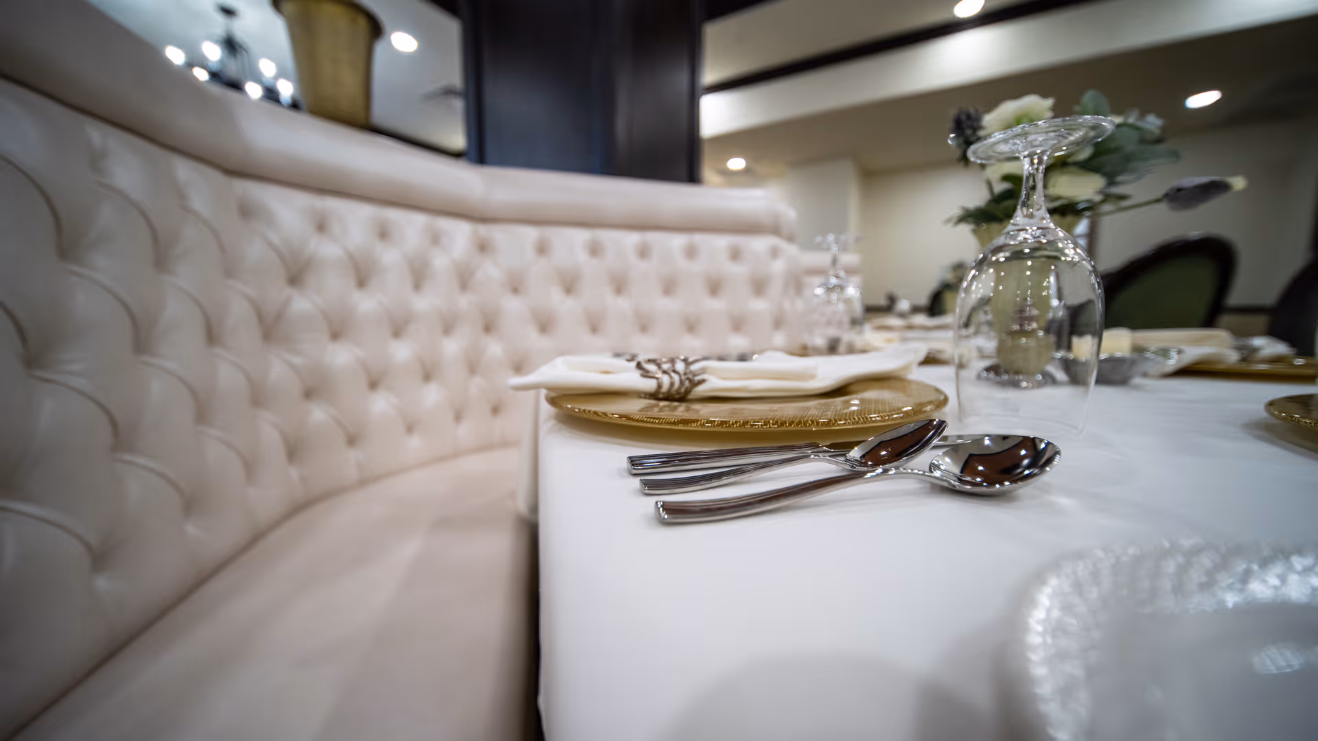 Close-up view of a dining table set with gold-rimmed plates, silverware, folded napkins, and upside-down wine glasses, next to a curved, tufted beige banquette seating in a softly lit dining area.
