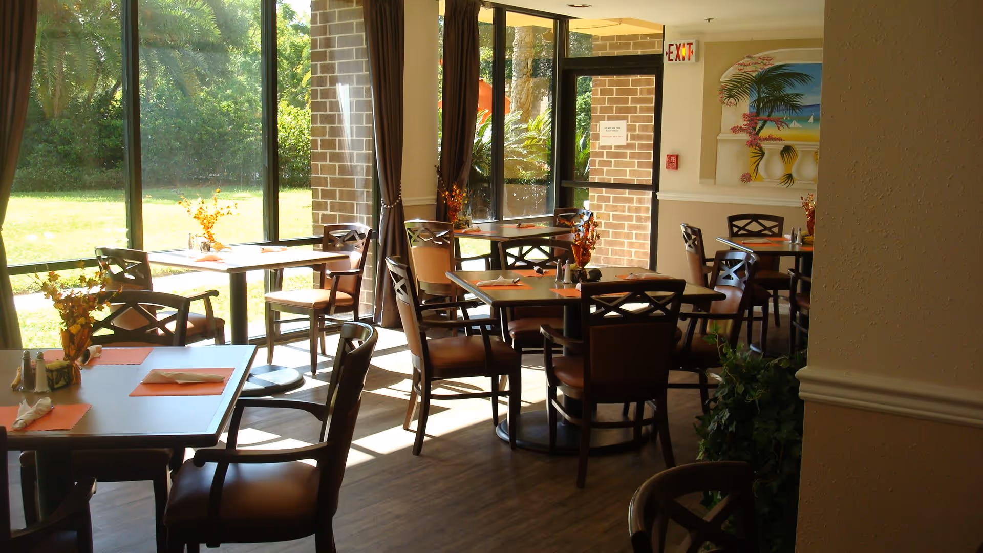 Bright dining room with multiple tables and chairs, large windows overlooking a grassy outdoor area and decorative plants.