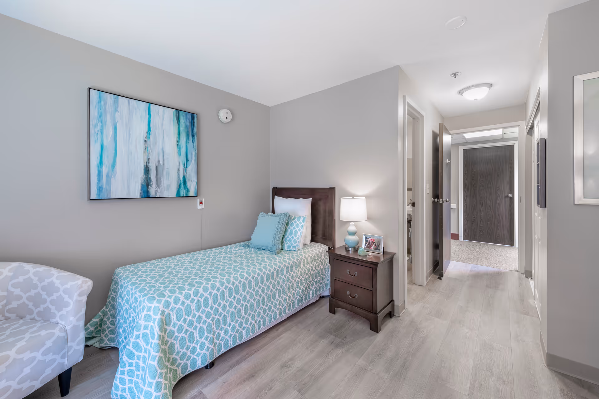 A neatly arranged bedroom in a senior living facility featuring a single bed with a turquoise patterned bedspread and matching pillows. Next to the bed is a dark wooden nightstand with a lamp and a framed photo. A light gray patterned armchair is positioned near the bed. The walls are painted light gray and decorated with abstract artwork. The room has light wood flooring and a hallway leading to other rooms.