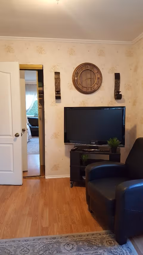 Small living room with a flat-screen TV on a black stand, a black armchair, a round wall clock on patterned wallpaper, and an open doorway to another room.
