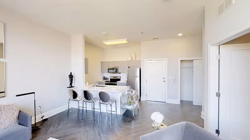 Bright and modern open-concept living space featuring a kitchen with stainless steel appliances including a refrigerator, microwave, and stove. The kitchen has a white island with three transparent bar stools. Adjacent to the kitchen is a cozy living area with a gray armchair and a small round table with a white flower arrangement. The room has light-colored walls, wood flooring, and recessed lighting. A doorway leads to a closet and another room.