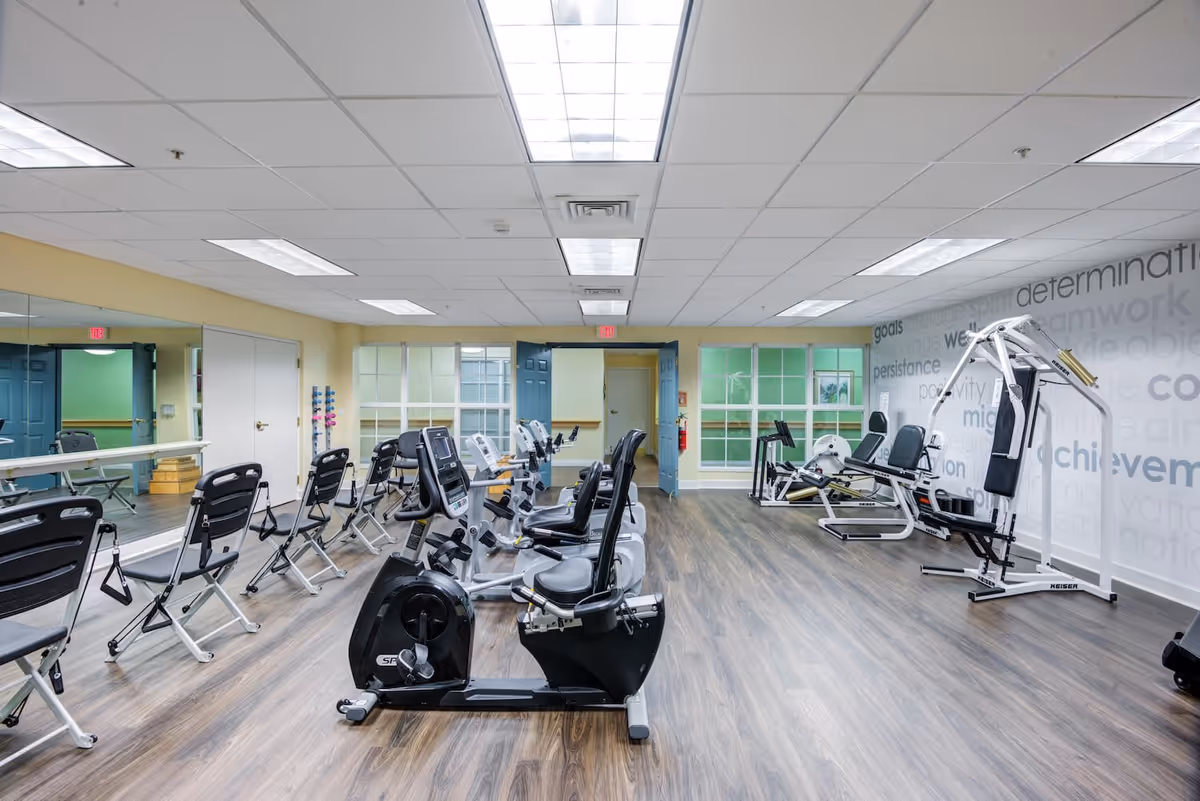A fitness room in a senior living facility with exercise bikes, weight machines, and folding chairs lined up along a mirrored wall. The room has wood flooring, bright overhead lighting, and motivational words on one wall.