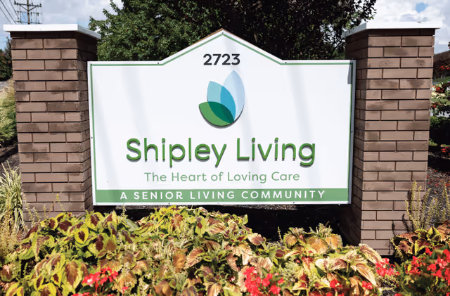 Outdoor sign for Shipley Living senior living community with the address number 2723, surrounded by plants and flowers under a partly cloudy sky.