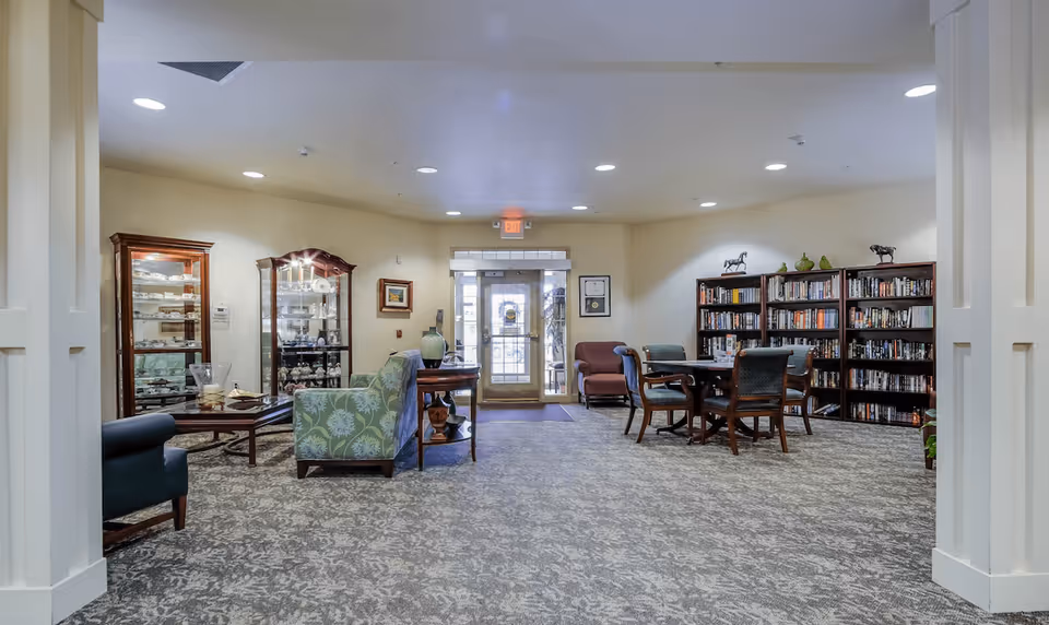 A spacious and well-lit common area in a retirement village featuring comfortable seating including armchairs and a sofa, a coffee table, display cabinets with decorative items, bookshelves filled with books, and a round table with chairs. The room has carpeted flooring, recessed ceiling lights, and a glass door leading outside.