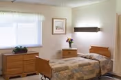 A simple, clean bedroom with a single hospital-style bed covered with a patterned quilt. There is a wooden dresser with a plant on top under a window with sheer curtains. A small wooden nightstand next to the bed holds a vase of flowers. A framed picture hangs on the wall above the nightstand, and a wall-mounted light fixture is above the bed.