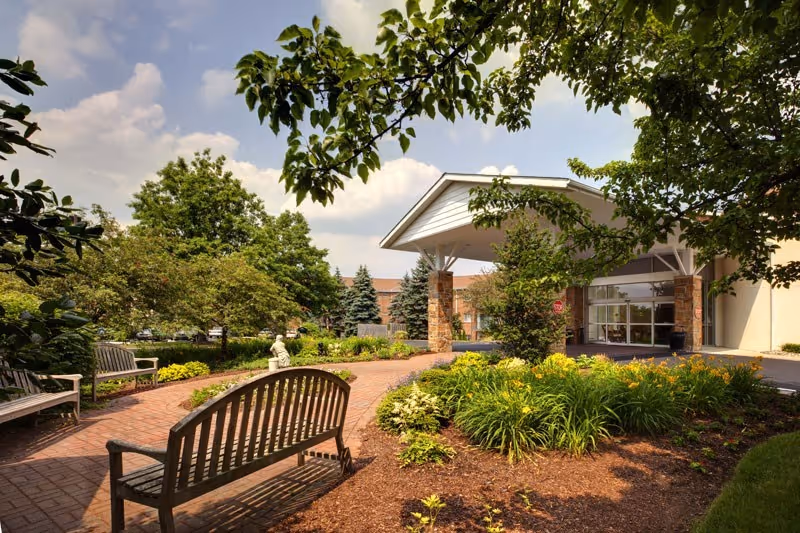 Outdoor garden area at Normandy Farms Estates featuring a brick pathway, wooden benches, lush green plants, trees, and a covered entrance to the building under a partly cloudy sky.