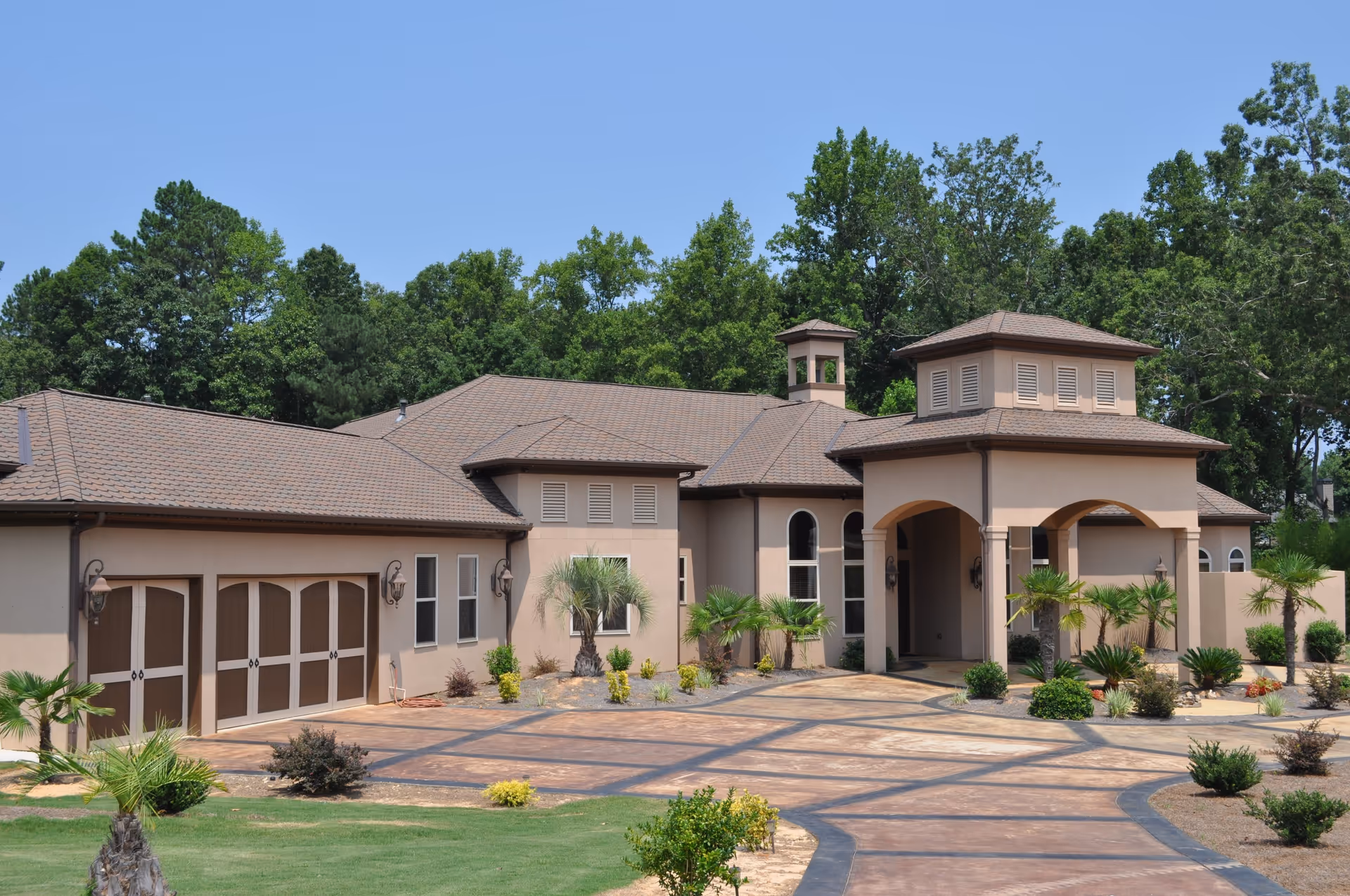 Exterior view of a single-story senior living facility building with a beige facade, brown roof, arched entrance, and surrounding landscaping including small palm trees and shrubs under a clear blue sky.
