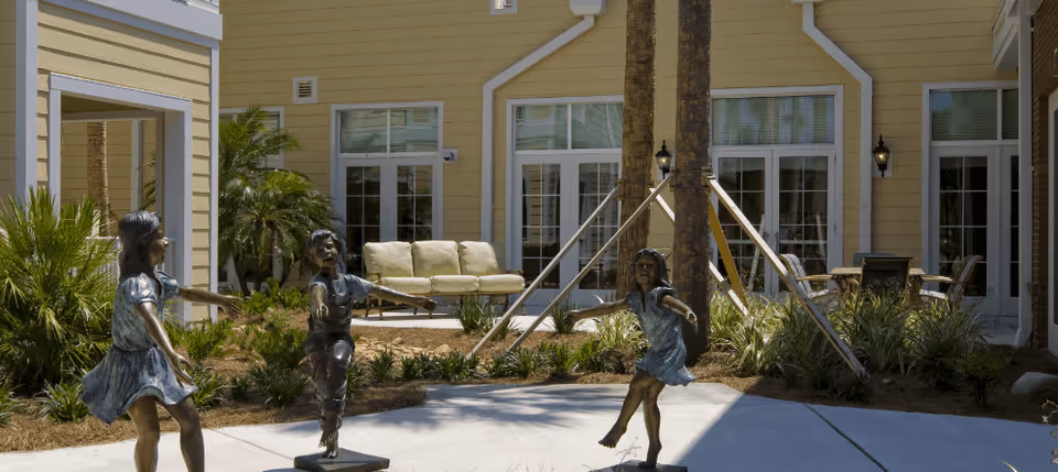 Bronze statues of three children dancing in a landscaped courtyard in front of a senior living building with patio furniture.