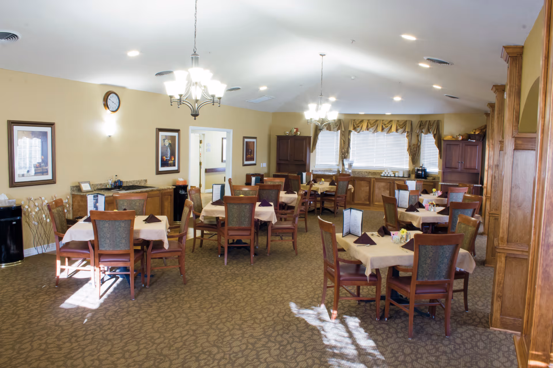 A well-lit dining room with multiple tables covered with beige tablecloths and set with menus and folded napkins. The room has carpeted floors, wooden chairs, chandeliers hanging from the ceiling, framed artwork on the walls, and large windows with curtains allowing natural light to enter. There are cabinets and a counter area along the walls.