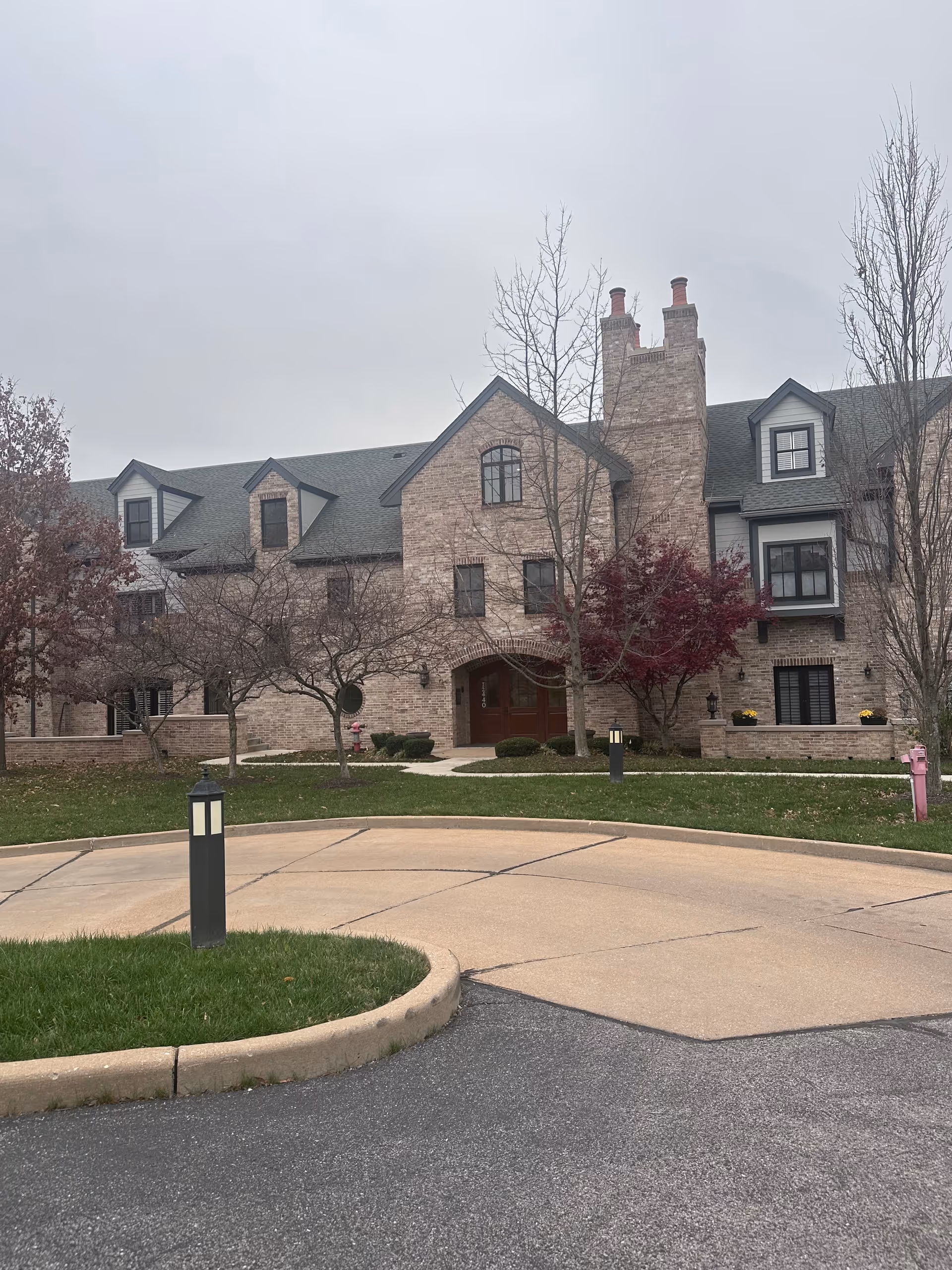 Exterior view of a large brick building with multiple windows and dormers, a central arched entrance, and two chimneys. The building is surrounded by a curved driveway, grass, and leafless trees with some red foliage. The sky is overcast.