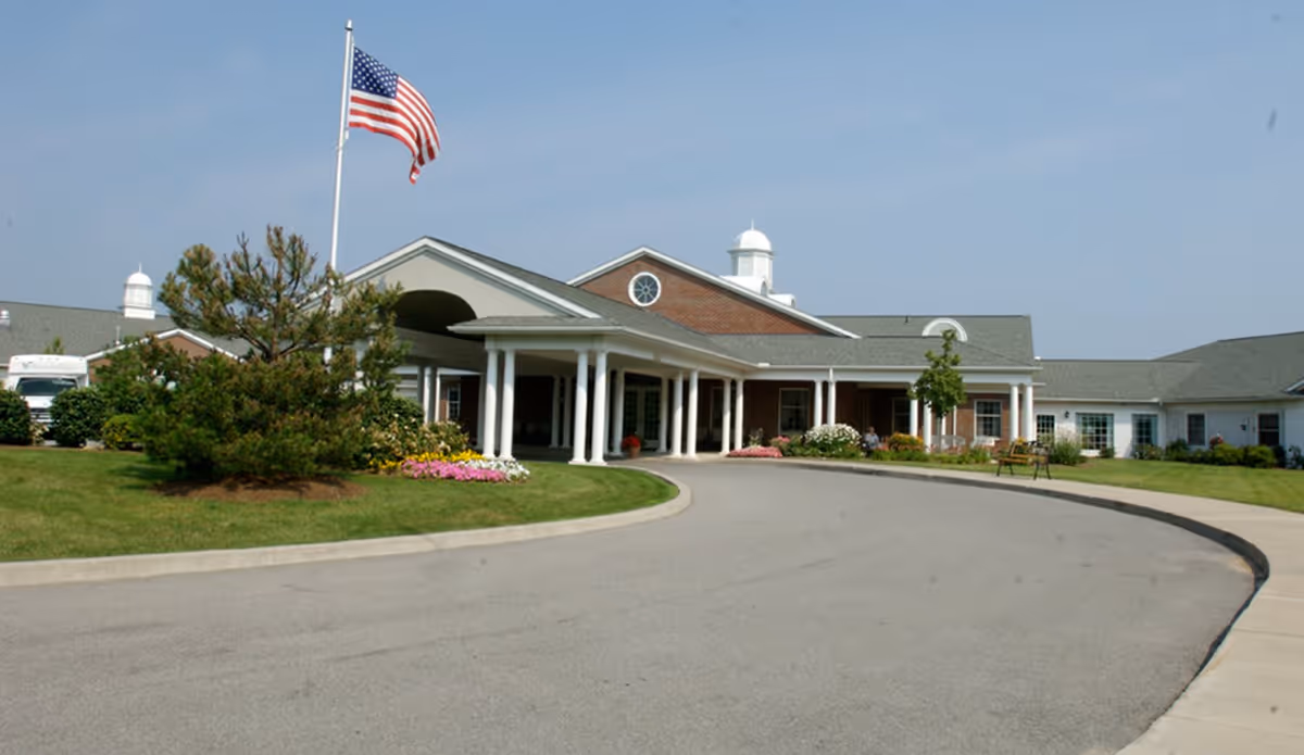 Exterior view of a single-story assisted living facility with a curved driveway, white columns at the entrance, an American flag on a flagpole, and landscaped greenery and flowers around the building.