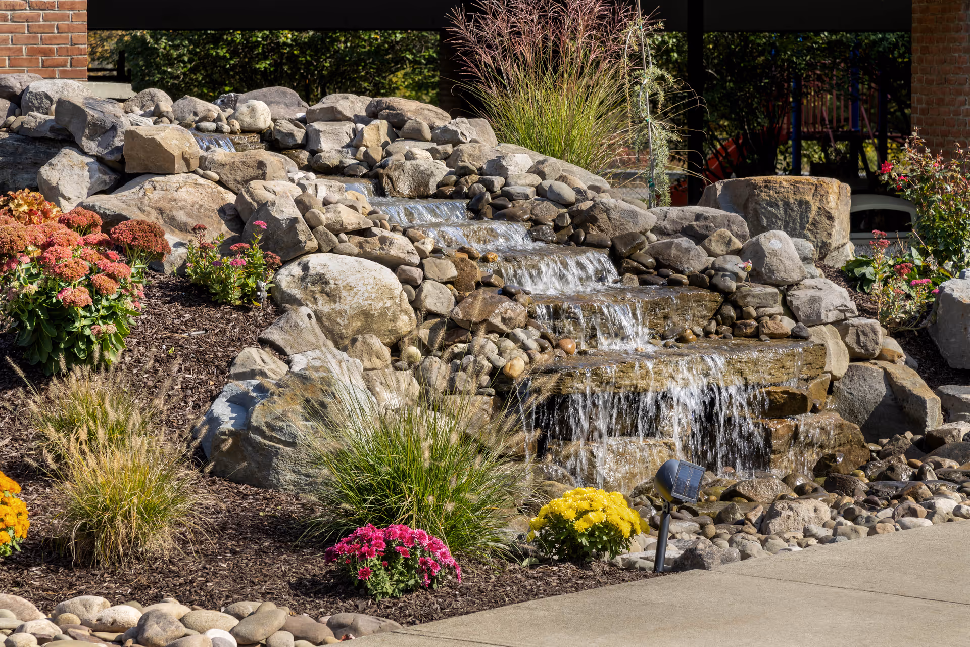A landscaped outdoor water feature with cascading water flowing over multiple levels of rocks surrounded by various flowering plants and ornamental grasses, with a brick building partially visible in the background.