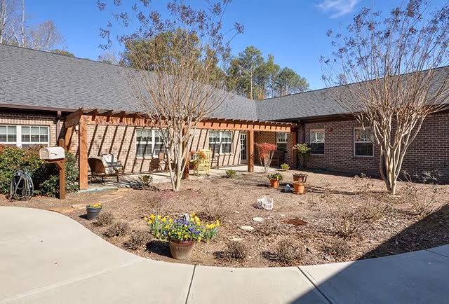 Outdoor courtyard area of a senior living facility with a brick building surrounding it. The courtyard features a wooden pergola with chairs underneath, several potted plants, leafless trees, and a concrete walkway curving around the garden beds. The sky is clear and blue.