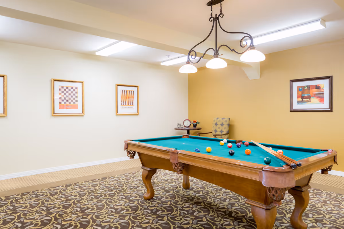 A well-lit room with a pool table in the center, featuring billiard balls and cues on the table. The room has patterned carpet flooring, beige and yellow walls adorned with framed artwork, a small round table with a clock and flower vase, and a cushioned chair in the corner. A decorative ceiling light fixture hangs above the pool table.