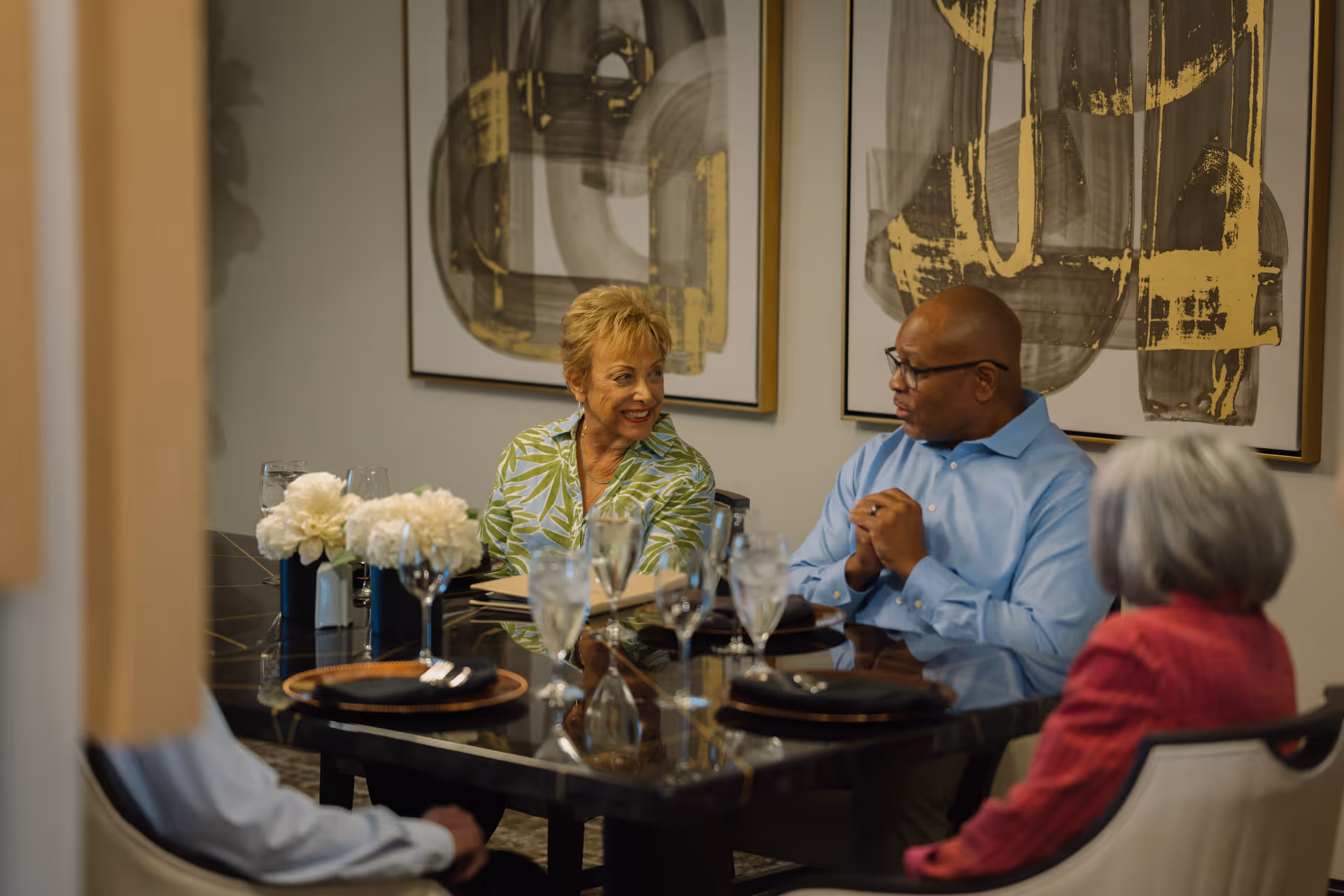 A small group of older adults seated around a dining table set with glasses, plates, and a floral centerpiece in a decorated dining room.
