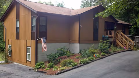 Brown two-story building with an American flag, exterior stairs, and landscaped slope beside a paved driveway.